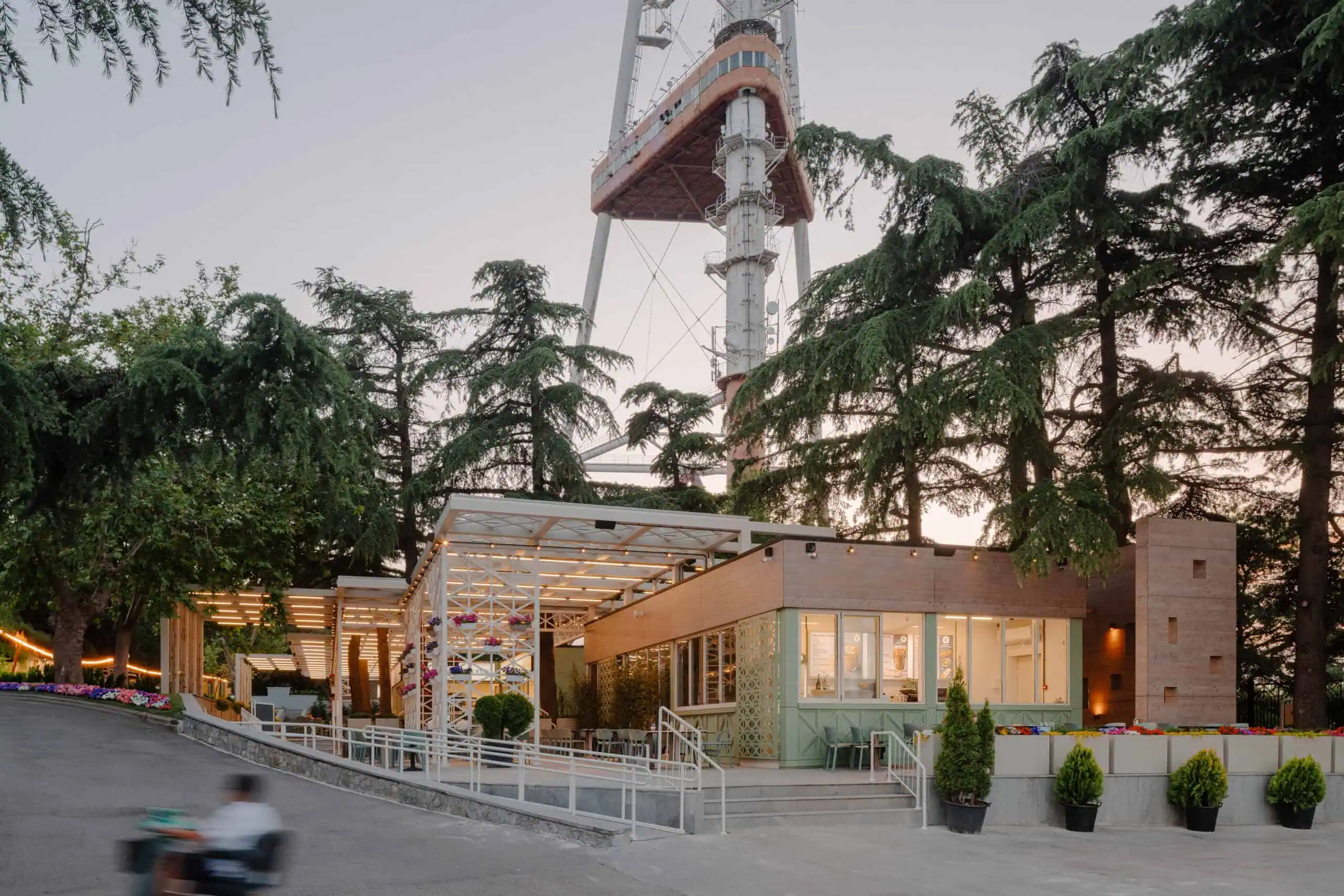 Modern restaurant with outdoor seating at sunset, surrounded by tall trees and city tower in the background.