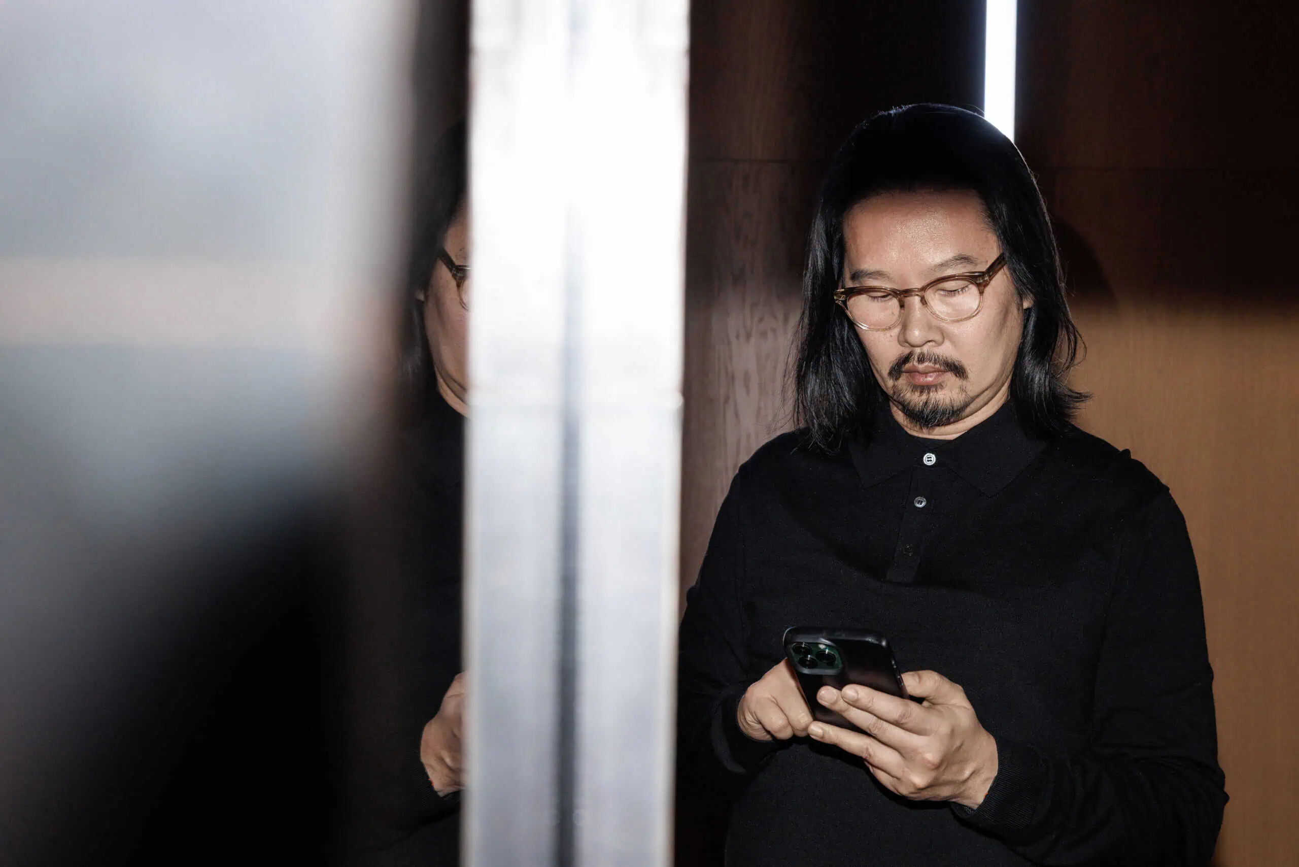 Modern man with long dark hair and glasses using a smartphone indoors at Studio 9.