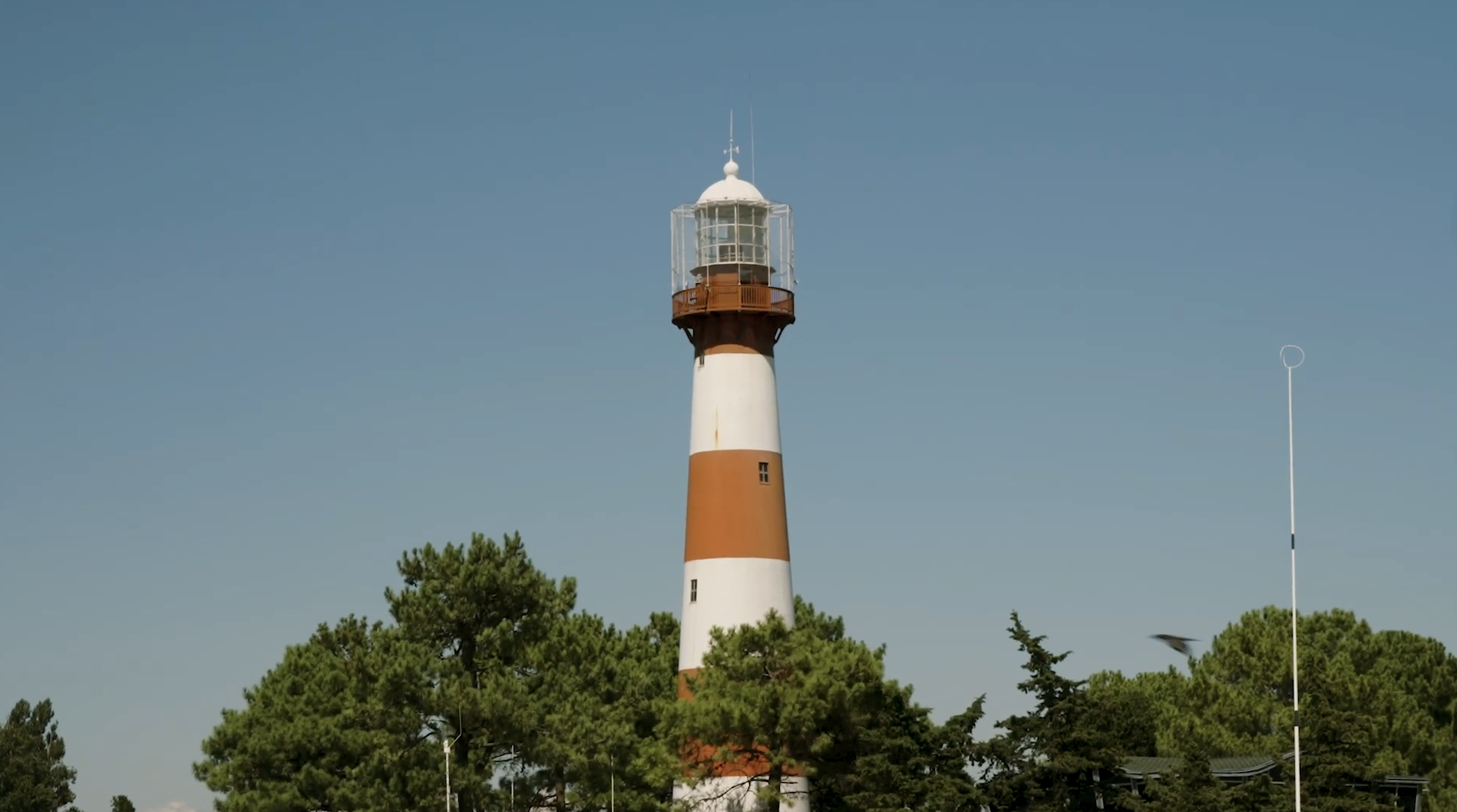 Historic lighthouse with orange and white stripes in scenic green surroundings under clear blue sky.